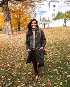 a woman standing in front of a tree with leaves on the ground and wearing a coat