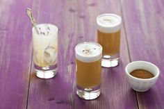 three glasses filled with drinks sitting on top of a purple wooden table next to two small bowls