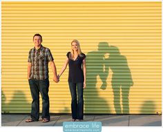 a man and woman holding hands standing in front of a yellow wall with shadows on it