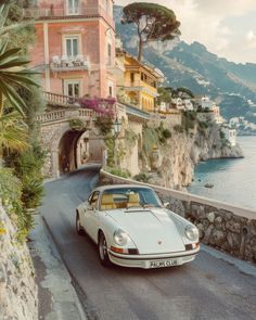 a white car driving down a road next to a cliff side with houses on it