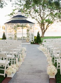 an outdoor ceremony setup with white chairs and flowers on the aisle, in front of a gazebo