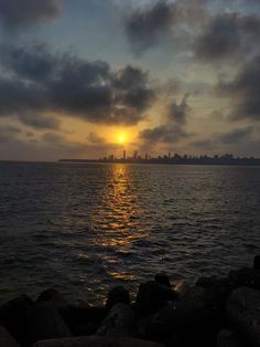 the sun is setting over the ocean with clouds in the sky and buildings on the horizon