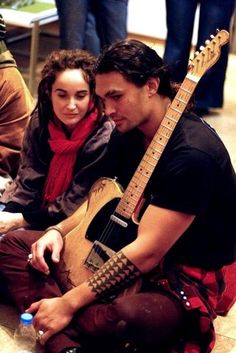 a man sitting on the ground with a guitar in his lap next to a woman