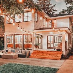 a large white house with lots of windows and lights on the front porch, surrounded by greenery