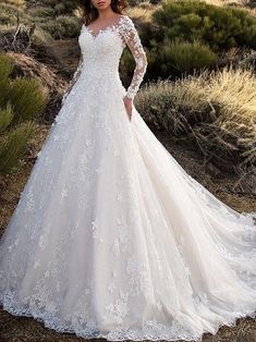 a woman in a white wedding dress standing on a dirt field with bushes and trees