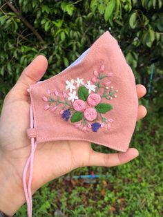 a hand holding a pink cloth face mask with flowers embroidered on the front and side