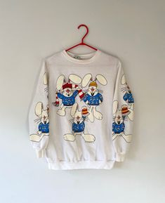 Rare! this colorful novelty pullover sweater from the 90s features an all over print of happy cartoon bunnies. there are three bunnies on the front of the sweater and two on each of the sleeves. the bunnies are each wearing blue sweaters with white bunnies on them, and some of the bunnies are also wearing toques and scarves, and the white paint of the bunnies are slightly puffy. the sweater has a white (slightly sheer) body, long sleeves and a crew neckline. the fabric is a soft cotton polyester Playful Crew Neck Sweater With Graphic Print, Playful Crew Neck Sweater With Cartoon Print, Fun Cartoon Print Sweatshirt, Vintage Sweatshirt With Cartoon Print, Multicolor Crew Neck Sweatshirt With Cartoon Print, Cartoon Animals Cute, Cartoon Bunnies, Three Bunnies, White Bunnies