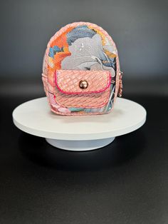 This mini clip-on backpack features a playful and stylish design. The primary material is textured in a woven basket-like pattern in soft pink, giving the bag a tactile and charming appearance. The front displays a mix of colors with abstract floral motifs in muted tones of blue, grey, orange, and yellow, adding an artistic flair. It has a small, practical front pocket with a button closure, detailed in bright pink stitching, further accentuating the bag's vibrant personality. Its compact size and convenient clip-on strap make it ideal for accessorizing, offering both functionality and a pop of unique style. Perfect for holding small essentials or attaching as a decorative accessory to larger bags. Matching Sling sold separately https://beralusvintondesign.etsy.com/listing/1807063053 size Tiny Bag, Small Pouch, Small Pouches, Coin Purses, Purse Pouch, Large Bag, Abstract Floral, Haiti, Handmade Shop