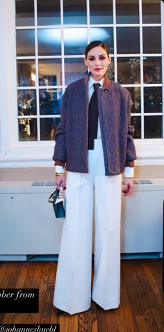 a woman standing in front of a window wearing white pants and a purple jacket with a black tie