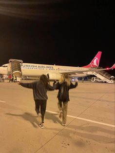 two women are walking towards an airplane on the tarmac at night with their arms in the air