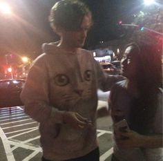 a man standing next to a woman on a street at night with a cat face drawn on his sweater