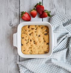 a white casserole dish with chocolate chip cookies and strawberries on the side