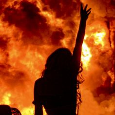 a woman reaching up to the sky with her arms raised in front of a huge fire