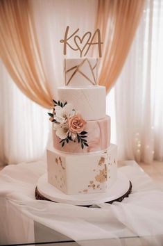 a three tiered wedding cake with pink flowers on the top and gold initials on the side