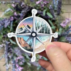 a hand holding a compass sticker with mountains and trees in the background, surrounded by purple flowers