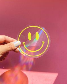 a person holding a smiley face sticker in front of a pink wall with the sun shining through it