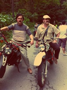 two men on motorbikes in the middle of a road with trees and bushes behind them