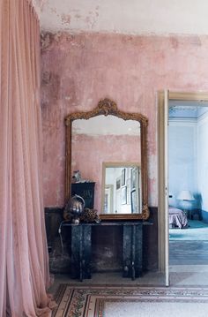 a mirror sitting on top of a wooden table next to a pink wall covered in curtains