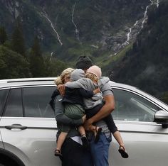 three people are hugging each other in front of a car with mountains in the background