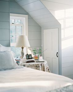 a white bed sitting under a window next to a table with a lamp on it