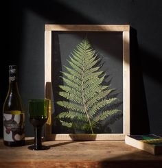 a green fern leaf in a frame next to a wine glass and bottle on a table