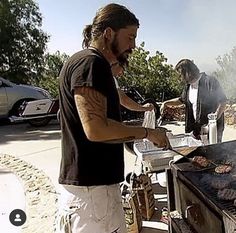 a man cooking food on top of a grill next to another man with long hair