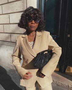 a woman in a tan suit and sunglasses standing on the street with her handbag