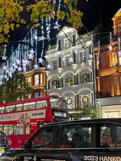 a red double decker bus driving down a street next to tall buildings covered in christmas lights