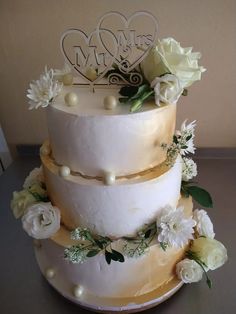 a three tiered wedding cake with white flowers and hearts on the top, sitting on a table