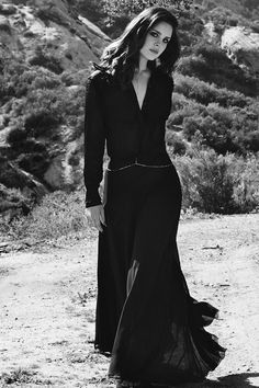 a woman in a long black dress is standing on the dirt road with her hands behind her back