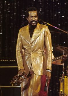 a man in a gold suit and red pants standing on stage with a microphone behind him