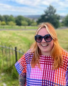 a woman with red hair and sunglasses standing in front of a fence smiling at the camera