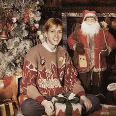 a man sitting in front of a christmas tree next to a santa clause and other decorations