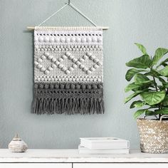 there is a plant and some books on the shelf in front of this crocheted wall hanging