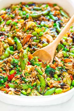 a white bowl filled with rice and vegetables next to a wooden spoon on top of a table