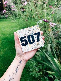 a person holding up a small square tile with the number 507 on it in front of some flowers