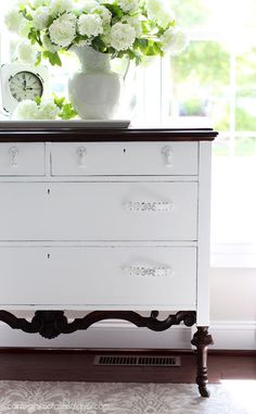 a white dresser with flowers and a clock on it's top, in front of a window