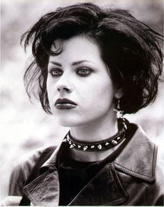 a black and white photo of a woman with short hair wearing a leather jacket, choker necklace and earrings