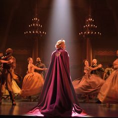 a woman in a purple cape and dress standing on stage with other dancers behind her