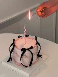 a person lighting a candle on top of a pink and white cake with black ribbons