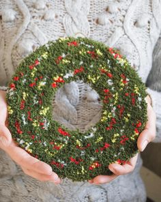 a person holding a green wreath with red and yellow berries on it in their hands