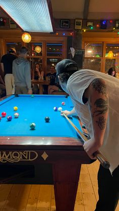 a man in white shirt and black pants playing pool with other people watching from the back