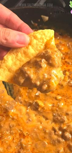 a hand holding a tortilla chip over a skillet filled with chili and cheese