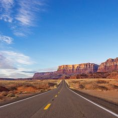 an empty road in the desert with mountains in the background and blue skies above it