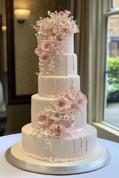 a three tiered wedding cake with pink flowers on the top and bottom, sitting on a table in front of a window