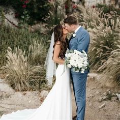 a bride and groom kissing in front of some tall grass with flowers on the ground