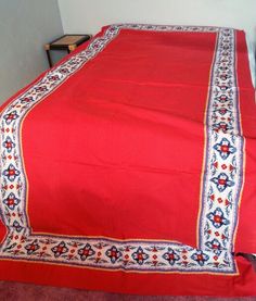 a red bed spread with white and blue designs on the border is sitting in front of a wall