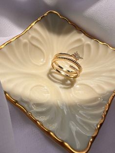 two gold wedding rings sitting on top of a white square plate in the shape of a flower