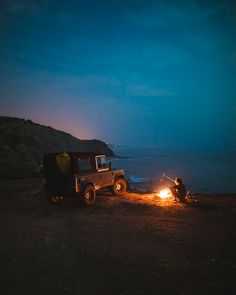 two people sitting around a campfire with a jeep in the background at night time