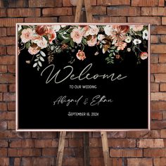 a welcome sign hanging on the side of a brick wall with flowers and leaves painted on it